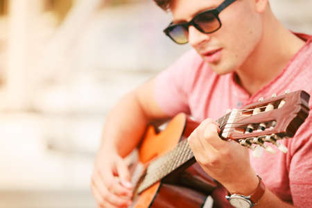 Performance and show time. Young fashionable man wearing sunglasses playing classic guitar outdoor.