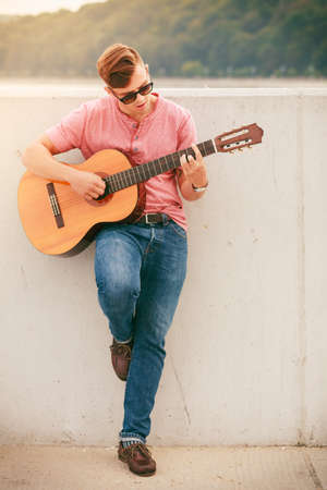 Performance and show time. Young fashionable man wearing sunglasses playing classic guitar outdoor.