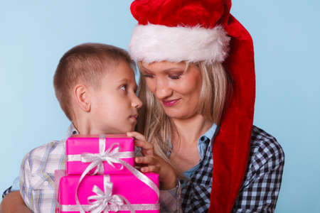 Happy childhood, holidays christmas time. Mother in santa claus hat and little boy child with stack of pink presents gift boxes on blueの写真素材