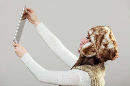 Technology and fun concept. Beauty young happy woman in warm winter clothes taking selfie photo self picture using tablet in studio on gray.の写真素材