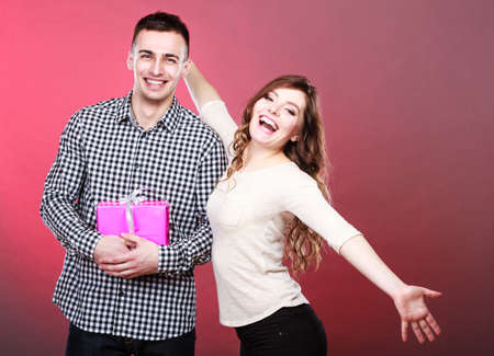 Couple and holidays concept. Handsome young man surprising lovely woman with pink gift box on red backgroundの写真素材