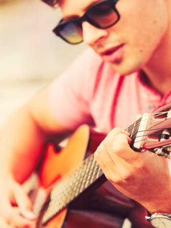 Performance and show time. Young fashionable man wearing sunglasses playing classic guitar outdoor.の写真素材
