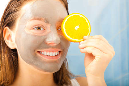 Beauty skin care cosmetics and health concept. Young woman with facial clay mask holding orange fruit slice covering eyes in bathroomの写真素材