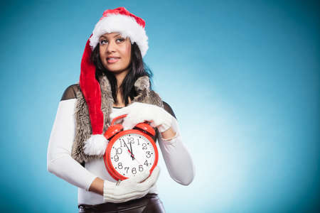 Happy cute woman in santa claus hat holding alarm clock. Attractive mixed race african girl in...