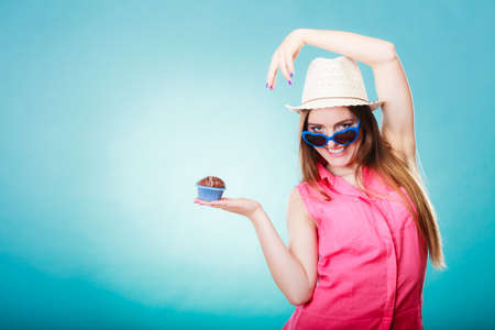 sweet food happiness and people concept. Smiling summer fashionable woman wearing straw hat heart shaped sunglasses holds cake cupcake in hand blue backgroundの写真素材