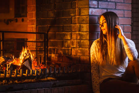 Melancholic pensive woman relaxing resting at fireplace. Thoughtful nostalgic young girl heating warming up. Winter at home.の写真素材