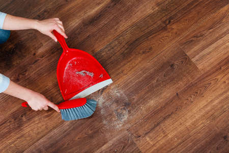Cleanup housework concept. Closeup cleaning woman sweeping wooden floor with red small whisk broom and dustpan indoorの写真素材