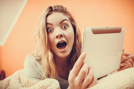 Amazed young woman using computer tablet browsing internet. Girl laying in bed under blanket and...
