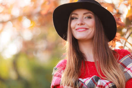 Portrait of attractive young smiling woman in autumnal park. Fashionable girl wearing red sweater scarf and black hat.の写真素材