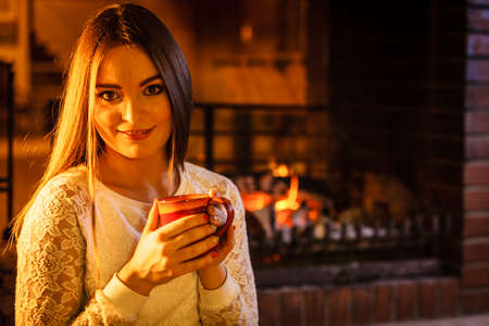 Woman drinking cup of coffee relaxing at fireplace. Young girl with hot beverage heating warming up....