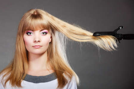Hairstyling. Attractive blonde woman long haired making hairstyle hairdo with electric hair iron straightener gray backgroundの写真素材