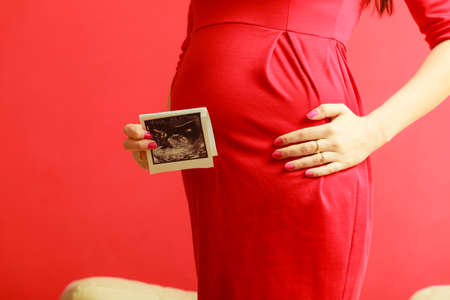 New life concept. Pregnancy, motherhood and happiness. Closeup tummy of pregnant woman wearing elegant red dress, ultrasound scan in hand indoorの写真素材