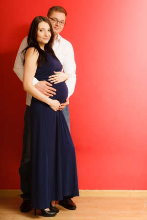 Happy family and waiting for baby concept. Handsome man husband hugging his pregnant attractive woman wife, portrait in full length on redの写真素材