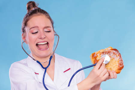 Scared terrified dietitian nutritionist checking examine sweet roll bun with stethoscope. Woman with fattening junk food. Bad unhealthy eating nutrition concept.の写真素材