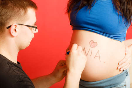 Pregnancy, parenthood and happiness concept. Family expecting new baby, having fun, man painting name of unborn baby girl writing on belly of pregnant womanの写真素材