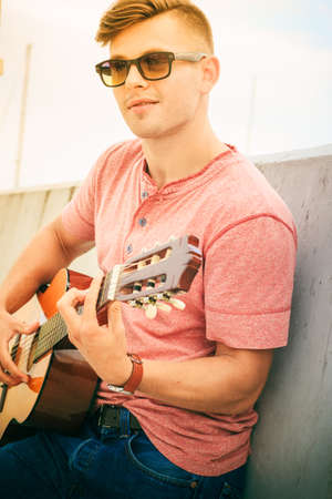 Performance and show on fresh air. Young fashionable man wearing sunglasses playing classic guitar outdoor. Summer time.の写真素材