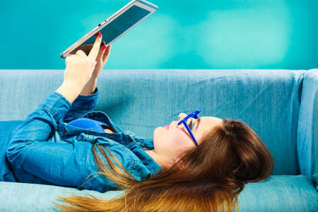 Modern technology education internet concept. Fashion young woman in glasses with tablet relaxing on blue color unusual viewの写真素材