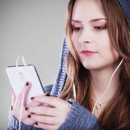 young woman with smart phone listening music. Teen stylish long hair girl in hood relaxing or learning language. Studio shot on gray.の写真素材