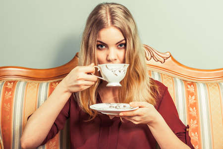 Fashionable woman drinking cup of coffee sitting on vintage sofa. Young girl with hot energizing beverage stay awake. Caffeine energy.の写真素材