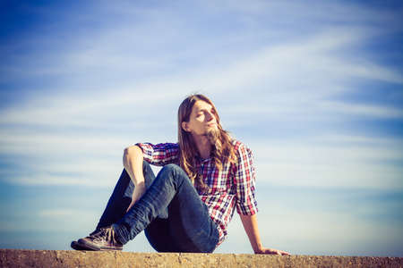 Man long hair wearing plaid shirt relaxing outdoor sitting on concrete wall at sunny windy day against blue skyの写真素材