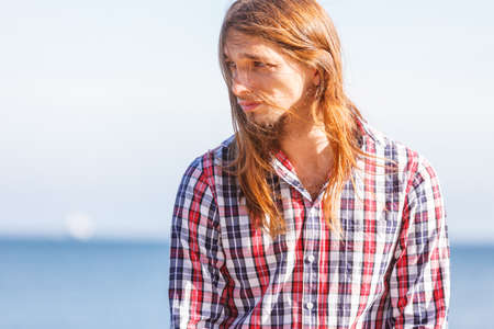 Man bearded long hair wearing plaid shirt casual style relaxing by seaside at summer sunny windy dayの写真素材