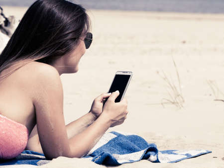 Communication concept. Young woman spending time on summer beach texting messages on smartphone. Girl using mobile phone.の写真素材