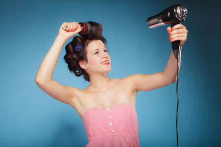 Young woman preparing to party having fun, funny girl styling hair with curlers hairbrush and hairdreyer retro style blue backgroundの写真素材