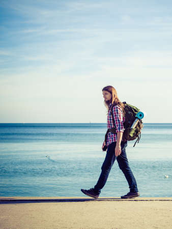 Man hiker backpacker walking with backpack by seaside at sunny day. Adventure, summer, tourism active lifestyle. Young long haired guy trampingの写真素材