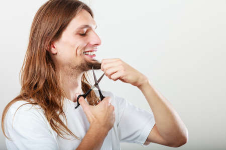 Cut and shave concept. Young man with long beard holding scissors. Boy cutting hair on chin.の写真素材