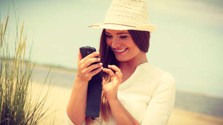 Communication concept. Young woman spending time on summer beach texting messages on smartphone. Girl using mobile phone.の写真素材