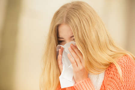Sick woman in fall autumn park sneezing in tissue. Ill girl caught cold flu outdoor. Rhinitis or allergy. Health care.の写真素材