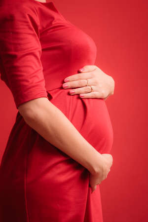 New life concept. Pregnancy, motherhood and happiness. Closeup on tummy of pregnant woman wearing stylish elegant red dress indoorの写真素材