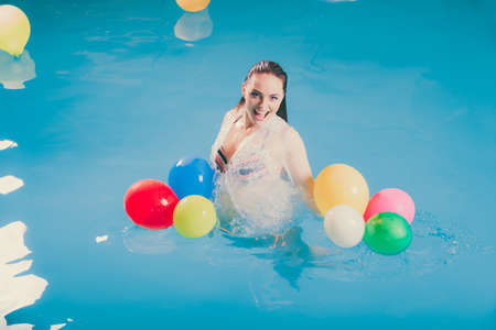 Happy woman in swimming pool water having fun with balloons. Seductive young girl wearing wet white...