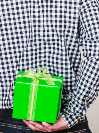 Man hiding green gift box with white ribbon behind back. Closeup of male hand holding christmas...