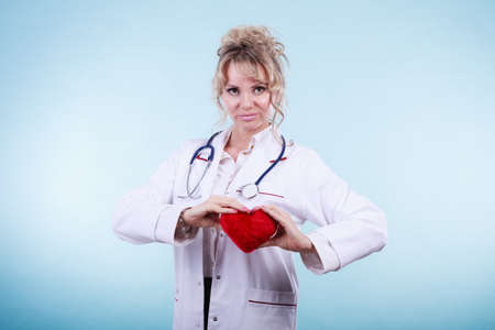 Love from working and helping people. Mid age blonde doctor in white medical apron with heart on blue. Enjoy work concept.の写真素材