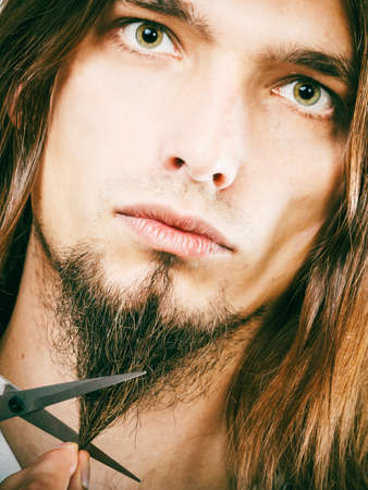 Cut and shave concept. Young man with long beard holding scissors. Boy cutting hair on chin.