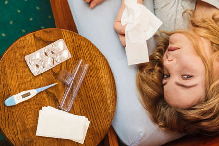 Sick woman laying in bed under wool blanket. Ill girl caught cold flu. Pills tablets and thermometer on table.の写真素材