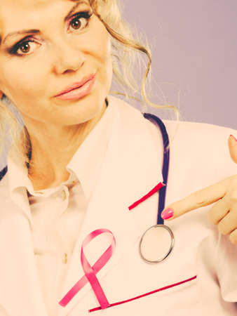 Breast cancer. Medical help treatment concept. Female blonde doctor in white medic apron showing pink ribbon and stethoscope. Filtered.の写真素材