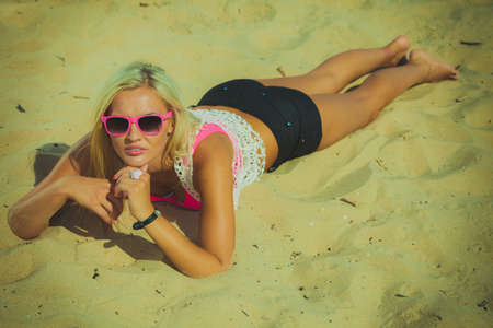 Pretty young blonde pretty girl at beach lying on sand. Woman wearing sunglasses have active time in summer. Summertime carefree concept.の写真素材