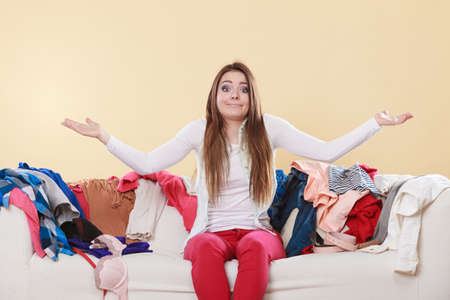 Helpless woman sitting on sofa couch in messy living room shrugging. Young girl surrounded by many stack of clothes. Disorder and mess at home.の写真素材