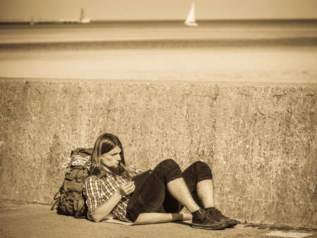 Man tourist backpacker relaxing outdoor sitting by grunge wall using tablet. Internet, tourism active lifestyle. Young hipster guy trampingの写真素材