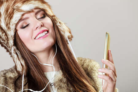 Winter girl listening music using phone with headphones. Happy woman wearing fur vest and warm hat in freezing cold time.の写真素材