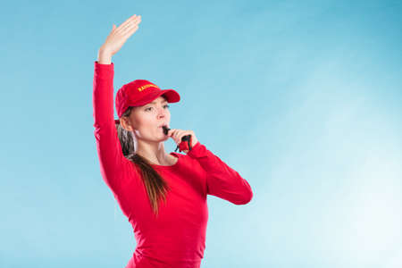 Lifeguard woman girl in red cap on duty supervising swimming pool water with raised arm hand blowing whistle on blue. Accident prevention rescue.の写真素材