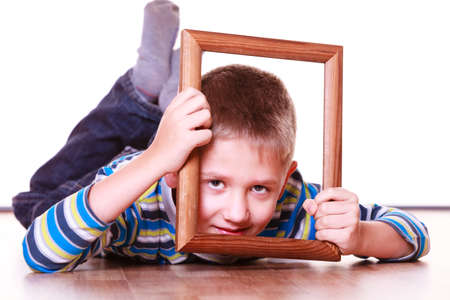 Spending free time and have fun. Playful little boy play with empty picture frame show face lie on floor.の写真素材