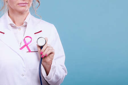 Women fight for health. Breast cancer tumor concept. Pink ribbon and blue stethoscope on white medical apron uniform. Doctor holding medic equipment.の写真素材