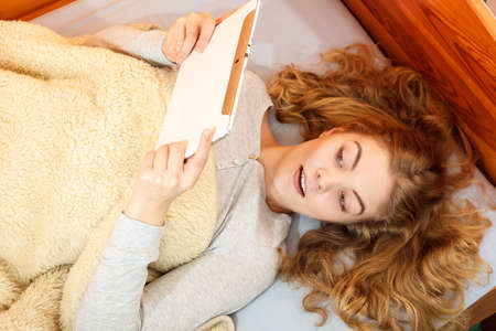 Lazy young woman using computer tablet browsing internet. Girl laying in bed under blanket and...