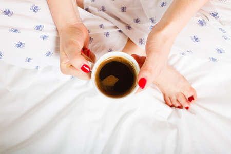 Woman relaxing at cozy home atmosphere on the bed. Female with red nails manicure holds cup of...