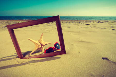 Summer photos concept. Frame on sand on the beach. Seashells and sunglasses lying on the seashore by the sea.の写真素材