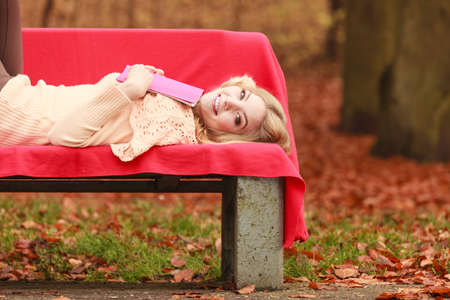 Happy smiling woman relaxing in fall park with book. Young blonde girl resting laying on bench. Autumn lifestyle fun.の写真素材