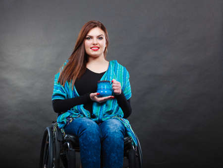 Real people, disability and handicap concept. Teen girl unrecognizable person sitting on wheelchair holding tea mug, studio shot on blackの写真素材